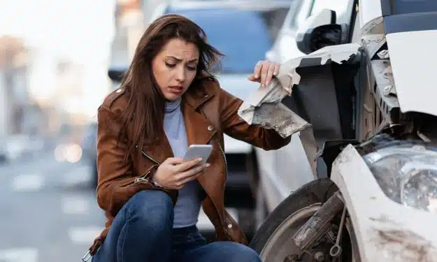 Woman squatting beside a damaged car while looking at her phone - car accident attorneys in Kennesaw, GA